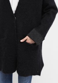Cappotto in lana nera con una superficie morbida e strutturata, con polsini in maglia grigia e chiusura a un bottone. Include tasche laterali.