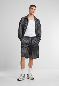 Veste de survêtement noire à rayures verticales accompagnée de shorts assortis. Le tissu semble lisse, avec une taille élastique et des poches latérales. Débardeur blanc et baskets.