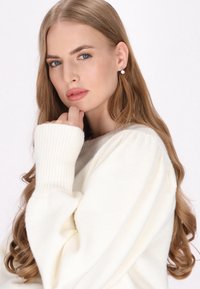 Sweter w kolorze kremowym, dziany, z prążkowanymi mankietami i okrągłym dekoltem. Długie faliste włosy, dyskretne kolczyki i delikatny, naturalny makijaż.