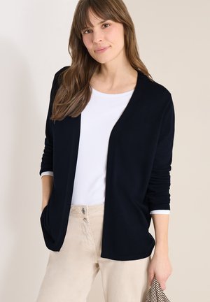 Jeune femme aux longs cheveux bruns portant un cardigan noir ouvert, une chemise blanche, un pantalon beige, tenant un tissu rayé dans une main.