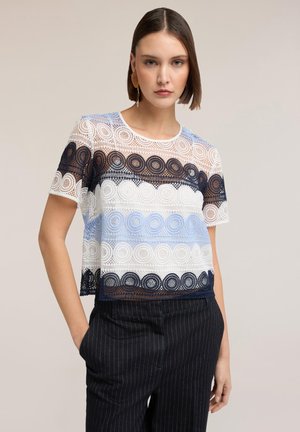 Młoda kobieta ubrana w krótkim rękawie koronkową bluzkę z okrągłymi wzorami w kolorach białym, granatowym i jasnoniebieskim, w parze z ciemnymi spodniami w prążki.
