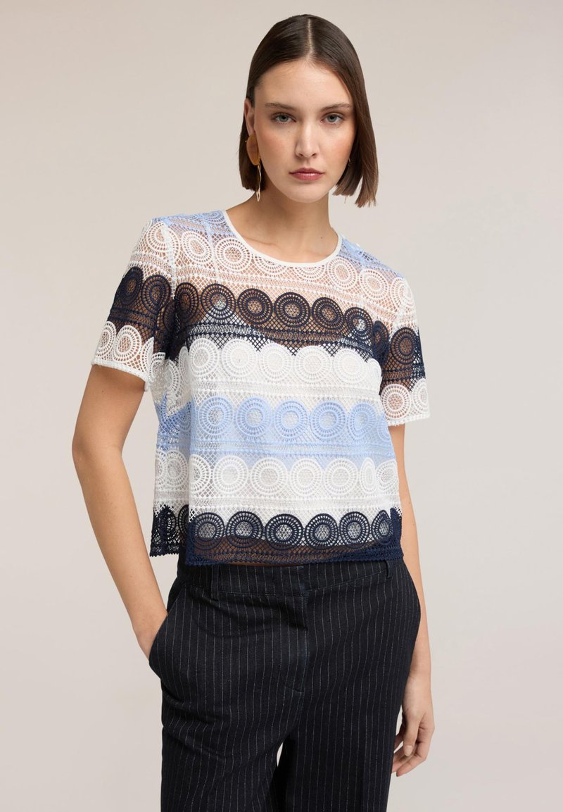 Młoda kobieta ubrana w krótkim rękawie koronkową bluzkę z okrągłymi wzorami w kolorach białym, granatowym i jasnoniebieskim, w parze z ciemnymi spodniami w prążki.