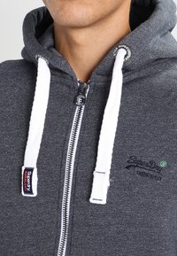Hombre que lleva una sudadera con cremallera en gris oscuro, con cordones blancos y el logo de Superdry bordado en el pecho.