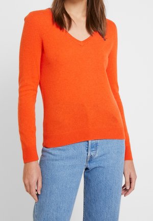 Femme portant un pull orange vif à col en V et un jean bleu clair, debout devant un fond clair uni.