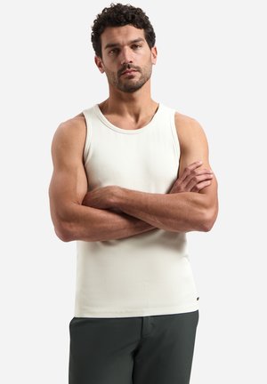 Man met kort gekruld haar die een wit, mouwloos geribd tanktopje en donkere broek draagt, met gekruiste armen tegen een witte achtergrond staat.