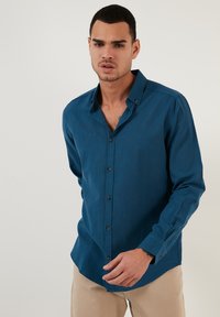 Teal lange-mouwen overhemd met donkere knopen, met een klassieke kraag en een gebogen zoom, gedragen met lichtbeige broek.