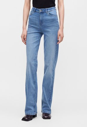 Persoon draagt lichtblauwe jeans met hoge taille en rechte pijpen, gecombineerd met zwarte schoenen met vierkante neus en een zwarte top, staand tegen een effen achtergrond.