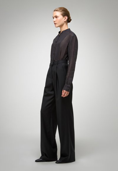 Chemise noire transparente à boutons associée à un pantalon large taille haute noir et des chaussures noires à bout pointu, mettant en valeur une silhouette élégante et structurée.