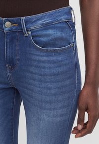 Gros plan d'une personne portant un jean denim bleu et un haut marron foncé, montrant la taille, la poche et une partie d'un bras et d'une main.