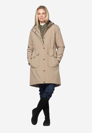 Woman wearing a beige knee-length coat over green shirt, blue jeans, and black boots, standing with hands in pockets and legs crossed.