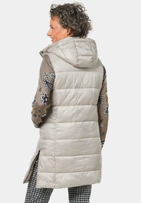 Beige gepolsterte Weste mit Kapuze, die ein gestepptes Design und kontrastierende gemusterte Ärmel aufweist. Seitenschlitze verbessern die Bewegungsfreiheit.