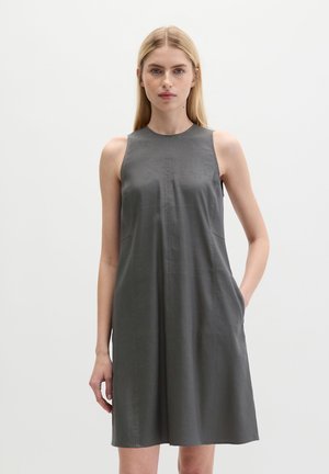 Kobieta w ciemnoszarej sukience bez rękawów do kolan stoi na jednolitym białym tle.