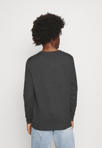 Persona con peinado afro, vistiendo una camiseta de manga larga gris oscuro y jeans azul claro, vista desde atrás sobre un fondo blanco.