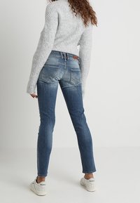 Jean bleu délavé avec une coupe droite et des accents en patch, associé à un pull côtelé gris clair et des baskets blanches.