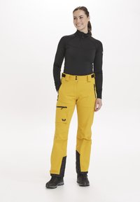 Gule skibukser med sorte detaljer, sidelomme med lynlås, justerbar talje og et slankt, vandtæt materiale. Båret sammen med en sort langærmet top.
