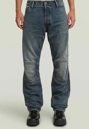 Jeans in denim blu scuro sbiadito con pannelli cuciti sulle ginocchia, indossati con scarpe nere robuste con lacci, mostrati dalla vita ai piedi.