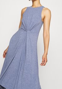 Robe bleue sans manches avec un détail froncé torsadé à la taille, portée par une personne avec un bras légèrement plié et l'autre détendu.