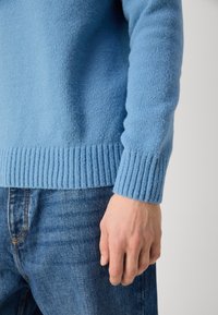 Main d'homme reposant à côté, portant un pull en tricot bleu clair et un jean en denim bleu sur un fond uni.