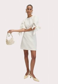 Krátké bílé šaty z texturované látky, nabírané rukávy s mašlemi a texturovaná taška přes rameno. Outfit doplňují neutrální boty na podpatku.