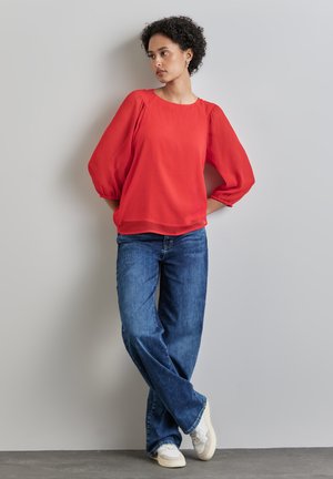 Rotes, locker sitzendes, strukturiertes Oberteil mit Ballonärmel, kombiniert mit blauen weit geschnittenen Jeans und weißen Sneakers. Einfacher runder Halsausschnitt, lässiger Stil.