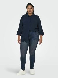 Blusa blu navy con bottoni e dettagli in pizzo sulle maniche, abbinata a jeans skinny in denim scuro e sneakers bianche. Tessuto morbido, stile casual.