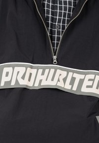 Vue partielle d'une veste noire zippée portée sur une chemise à carreaux noir et blanc, avec "PROHURTLTE" écrit sur la poitrine.