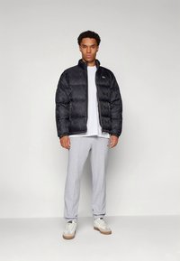 Juoda pūkinė striukė su sulenktu apykakliu, ant krūtinės – mažas logotipas. Derinama su šviesiai pilkomis sportinėmis kelnėmis ir baltais sportbačiais.