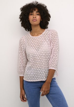 Top in pizzo rosa chiaro con un motivo geometrico, maniche a tre quarti e scollo rotondo. Abbinato a jeans blu.