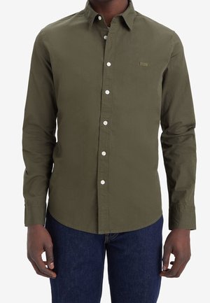 Mężczyzna w oliwkowozielonej, długorękawowej koszuli zapinanej na białe guziki i ciemnoniebieskich dżinsach, stojący na białym tle.