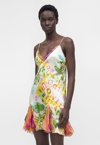 Γυναίκα που φοράει ένα πολύχρωμο φόρεμα με λουλούδια, λεπτές τιράντες και βολάν στο τελείωμα, στέκεται μπροστά σε απλό λευκό φόντο.