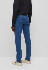 Jeans de mezclilla en azul oscuro con un corte recto, cinco bolsillos y detalles desgastados. Combinados con zapatillas beige con un acento en azul marino.