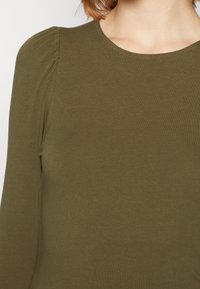 Frau, die ein olivgrünes Langarmoberteil mit Rundhalsausschnitt und dezenten Raffungselementen an der Schulter trägt.