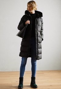 Manteau long noir en duvet avec une capuche, fermeture éclair, et texture matelassée, associé à un jean bleu et des bottes noires teintées de vert. Sac à main noir.