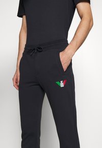 Black jogger pants made of soft fabric with a drawstring waist and side pockets. Features a colorful striped logo on the left thigh.