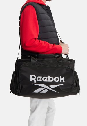 Osoba w czerwonej bluzie z kapturem i czarnej kamizelce niesie dużą czarną torbę sportową Reebok z paskiem na ramię i bocznymi kieszeniami.