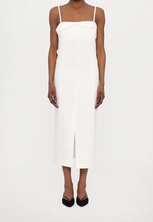 Frau trägt ein weißes Midikleid mit schmalen Trägern und einem vorderen Schlitz, kombiniert mit schwarzen spitzen Pumps, stehend vor einem einfarbigen Hintergrund.