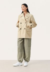 Beige dobbeltrækket jakke med store sorte knapper, sidelommer og en afslappet pasform, båret over olivenfarvede cargo-bukser og hvide sneakers.