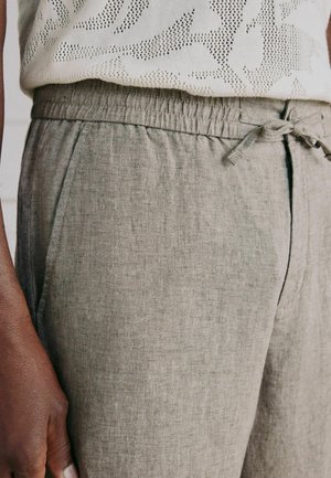 Prim-plan cu o persoană cu piele închisă la culoare purtând pantaloni din in bej cu talie elastică și șnur, asortați cu un top alb tricotat.