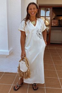 Abito bianco oversize a V con stampa grafica nera, indossato con una borsa di paglia e infradito con decorazioni.