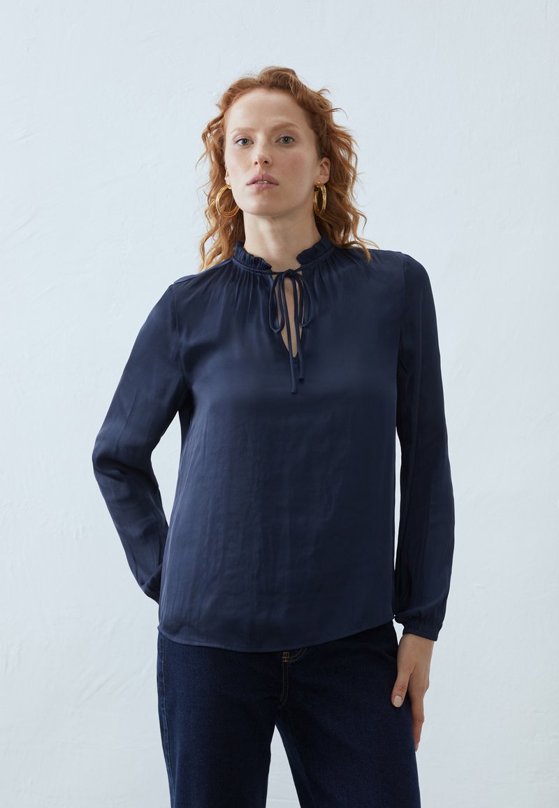 Blusa blu navy con maniche lunghe, scollo arricciato con chiusura a lacci, tessuto morbido e vestibilità rilassata, abbinata a jeans in denim scuro.
