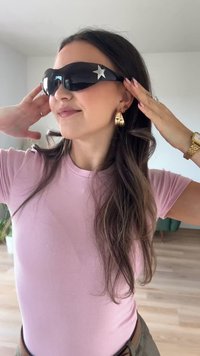 Μαύρα γυαλιά ηλίου με γυαλιστερή επιφάνεια και ασημένια διακόσμηση στην μορφή αστέρα, συνδυασμένα με χρυσές φόρμες σκουλαρίκια. Το μοντέλο έχει μακριά μαλλιά και φοράει ροζ μπλούζα.