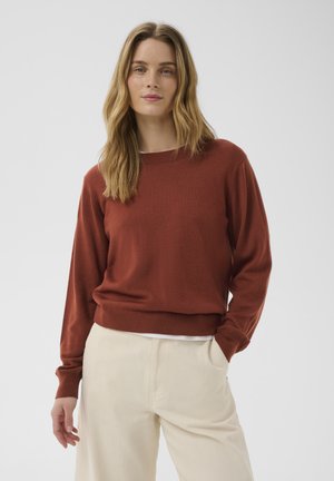 Femme aux longs cheveux blonds portant un pull couleur rouille et un pantalon crème, debout avec une main dans la poche devant un fond uni.