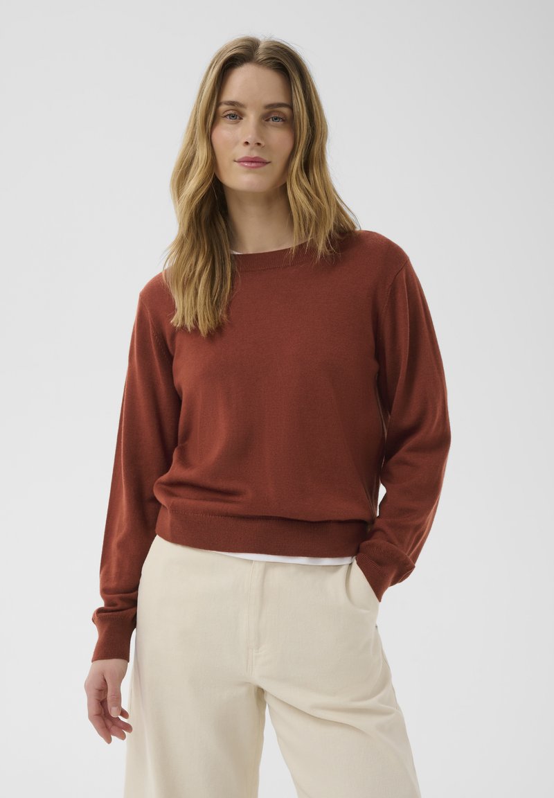Femme aux longs cheveux blonds portant un pull couleur rouille et un pantalon crème, debout avec une main dans la poche devant un fond uni.