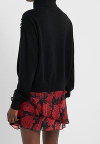 Schwarzer Rollkragenpullover mit lockerer Passform, ausgestattet mit strukturierten Details an den Schultern. Kombiniert mit einem gestuften roten Rock mit Blumengewinde.