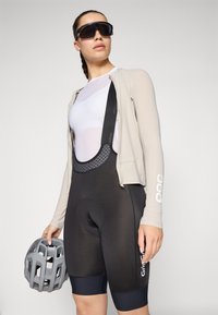 Sykkeldrakt med en lys beige langermet jakke, hvit mesh-skjorte og svarte polstrede bib-shorts, med en grå hjelm i hånden.