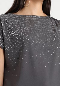 Šedý krátký rukáv s kulatým výstřihem, který zdobí gradient stříbrných teček na přední straně pro texturu a vizuální zajímavost.