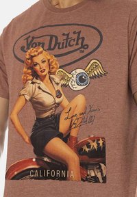 T-shirt en coton marron avec un graphique vintage représentant une femme en chemise blanche et jupe bleue, avec un œil et des ailes, étiqueté "California".
