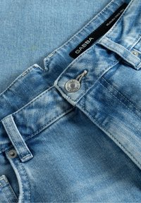 Jeans de denim azul claro con un lavado desgastado, que presentan un botón plateado, costuras en contraste y una etiqueta de marca negra cosida en la cinturilla.