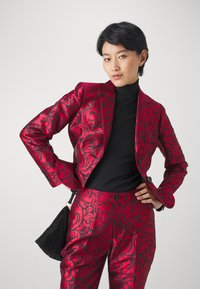 Punainen kukkakuosiset kankaat puku kiiltävästä materiaalista, yhdistetty mustan turtleneck-puseron ja mustan teksturoidun käsilaukun kanssa. Muotoiltu istuvuus.