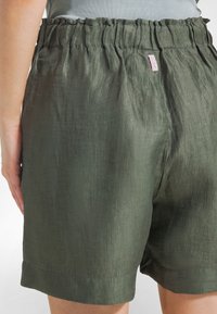 Pantaloncini verdi in lino con fascia elastica arricciata in vita, orlo arrotolato e dettaglio di etichetta rosa minimalista sul retro. Tessuto leggero e strutturato.